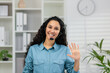 A cheerful woman in a blue shirt and headset waves to the camera, portraying a welcoming online customer service environment in a modern office.
