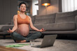 A focused mid adult pregnant lady meditating in her living room in front of a laptop