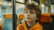 A boy with curly hair sits on a bus and looks out the window thoughtfully