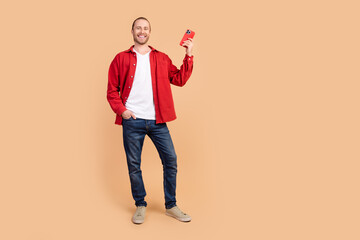 Wall Mural - Full length portrait of nice young man show hold smart phone wear red shirt isolated on beige color background