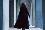 Fototapeta  - Young woman wearing long black cloak with red hair walking with her back turned in corridor between the columns of architectural building