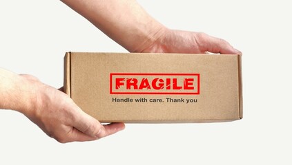 One rectangular cardboard box in hands for fragile items packing on a plain light background. An adhesive red warning label, sticker for packaging 'Fragile, Handle with care, Thank you'