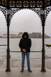 middle aged man wearing a black windbreaker posing under a gazebo by the lake on a rainy day - travel and tourism concept