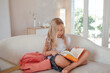 a little girl 6-7 years old is getting ready for school, the girl is dressed in a light-colored top and a plaid skirt.  The kid lsit on the sofa  with a backpack and a book