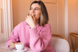 Happy woman eating cookie sitting in a restaurant. Positive woman bites cookie enjoys eating sweet food breaks diet looks away. Woman wear pink sweater rest in cafe. Girl bite cake and look happy.