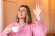 Attractive cheerful female adult gesture hand wave to say hi to her friend walking in cafe restaurant, happy woman hand hold coffee cup relax casual greeting to her friend in cafe.