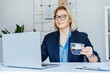 © okrasiuk - Smiling mature businesswoman working on laptop and drinking coffee at office, confident middle-aged experienced senior female professional, CEO of company. Female entrepreneur manage business.