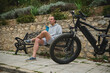 © Taras Grebinets - young man cyclist in sportswear, drinking water from sports bottle in the city, sitting on a bench, relaxing after riding an eco-friendly e-bike motorbike. Electric bicycle in the foreground