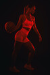 A beautiful athletic blonde girl in white shorts and a top plays basketball on a black background in red light.