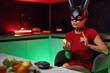 beautiful girl in a bdsm rabbit mask and a bright red dress eats an apple in the kitchen in neon light promoting a healthy lifestyle vegetarianism