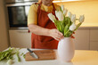 Happy Caucasian 30s woman taking care of cut fresh bouquet of tulips in kitchen. Housewife hands of woman cutting flower on wooden board and putting fresh tulips into the vase.