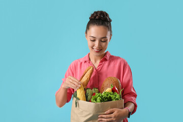 Poster - Beautiful young Asian woman holding paper bag with fresh products on blue background