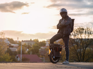 Wall Mural - A young male rider in protective gear with a backpack on an electric unicycle against the background of the city at sunset. Driving around the city on a mono wheel