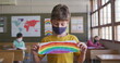 A Caucasian child in a yellow shirt and mask shows a rainbow drawing at school