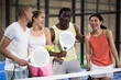 Happy laughing men and women of different nationalities in sportswear with rackets and balls in hands talking friendly near net on indoor padel court.