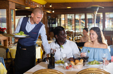 Wall Mural - Waiter serving meals for positive interracial couple at restaurant, putting plates with food on table