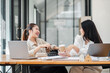 © Satori Studio - Two joyful businesswomen laughing and chatting over a cup of coffee at a wooden office desk with laptops.