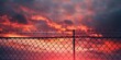 A chain link fence with barbed wire at sunset symbolizes security and protection, set against a dark red sky with storm clouds gathering above.