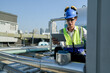 © ultramansk - Young technician with hard hat and safety vest adjusts equipment on a commercial building's rooftop amidst an urban landscape under a clear sky.