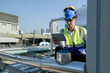 © ultramansk - Young technician with hard hat and safety vest adjusts equipment on a commercial building's rooftop amidst an urban landscape under a clear sky.