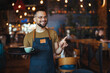 Portrait of smiling young waiter holding tray in cafe. Handsome waiter smiling at camera holding tray at the coffee shop. Profesional Waiter in Restaurant
