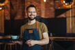 Happy confident young waiter entrepreneur looking at camera, smiling male small cafe business owner employee standing in restaurant, millennial businessman wear apron posing in coffee shop
