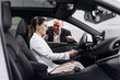 © Vladislav - Satisfied happy caucasian female client customer woman sitting at the wheel of new car doing test-drive before buying auto while male shop assistant helping her choose it
