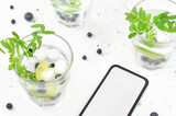 Fototapeta Kwiaty - Drinks in glasses and phone with empty space to fill with content. Decorations: leaves and blueberries on light table with little blue paint stains.