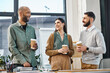 © Bliss - A group of coworkers standing around a table, engaging in a discussion over cups of coffee during a break in the office.