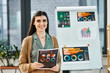 © Bliss - A woman stands by a whiteboard holding charts. She appears to be leading a presentation in a corporate office setting.