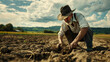 A rural man in a hat sat down in a muddy field. The farmer looks down, studying the soil and inspecting his plantings.