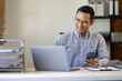 © Kainnika - Happy businessman in office using mobile phone to work with laptop on desk.