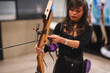 A beginner Asian woman is engrossed in learning archery, trying her hand at this challenging sport in an indoor range at a mall.
