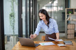 © Wasana - Smiling Asian businesswoman working at her desk with laptop, surrounded by minimalist style shelving in a contemporary office.