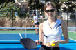© Mat Hayward - Female pickleball player wearing black and white sports shirt and sunglasses holding ball and paddle while standing outside during a match on a blue court.