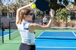 © Mat Hayward - Female pickleball player wearing black and white sports shirt and sunglasses holding ball and paddle while standing outside during a match on a blue court.