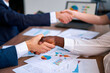 Multiracial business professionals seal agreement, shaking hands over data charts, teamwork, negotiation. Office setting, multiethnic inclusion in deal-making, signifies corporate unity, consensus.