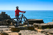 Brave senior woman riding her electric mountain bike on the rocky cliffs of Peniche at the western atlantic coast of Portugal