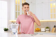 Happy man drinking clear water near filter jug at table in kitchen