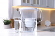 Water filter jug and glass on white marble table in kitchen, closeup
