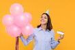 Happy young woman in party hat with balloons and cheesecake on yellow background