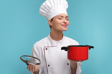 Wall Mural - Happy woman chef in uniform holding cooking pot on light blue background