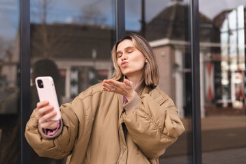 Poster - Happy woman using smartphone at cafe. Smiling blonde woman using smart phone make selfie, spends her leisure time on street. Blonde girl wear bomber. Girl send blow kiss to phone camera.