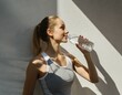 © Monika - Młoda kobieta w sportowym stroju oświetlona promieniami słońca stoi na tle ściany trzymając w ręku butelkę z wodą. Lato, sport, wakacje, zdrowy styl życia