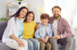 © Studio Romantic - Smiling young happy family of four with two kids boy and girl sitting on the sofa at home and looking cheerful at camera. Portrait of parents with children. Family time and leisure concept.