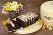 Chocolate chip cake with slices and icing drizzle on metal tray with lid on rustic wood background with Spring flowers. Homemade sweet food with floral arrangement.