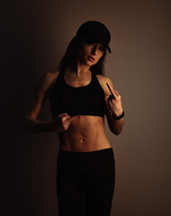 Wall Mural - Fuck you. Sport sexy muscular woman posing in black sport bra, cap and showing the fuck sign the hand, standing on dark shadow studio background. Front body view.