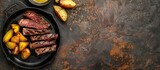 Fototapeta  - Cast iron plate holding grilled rib-eye steak slices, accompanied by yellow potato wedges and oil in a small dish, placed on a table with empty space for text.