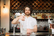A skilled male bartender with a beard mixes a cocktail in a stylish bar setup, showcasing expertise and a warm, inviting atmosphere.