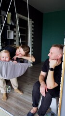 Sticker - Mother sways her lovely blond baby in a swing at home. Father sits beside watching his child smiling. Vertical video.